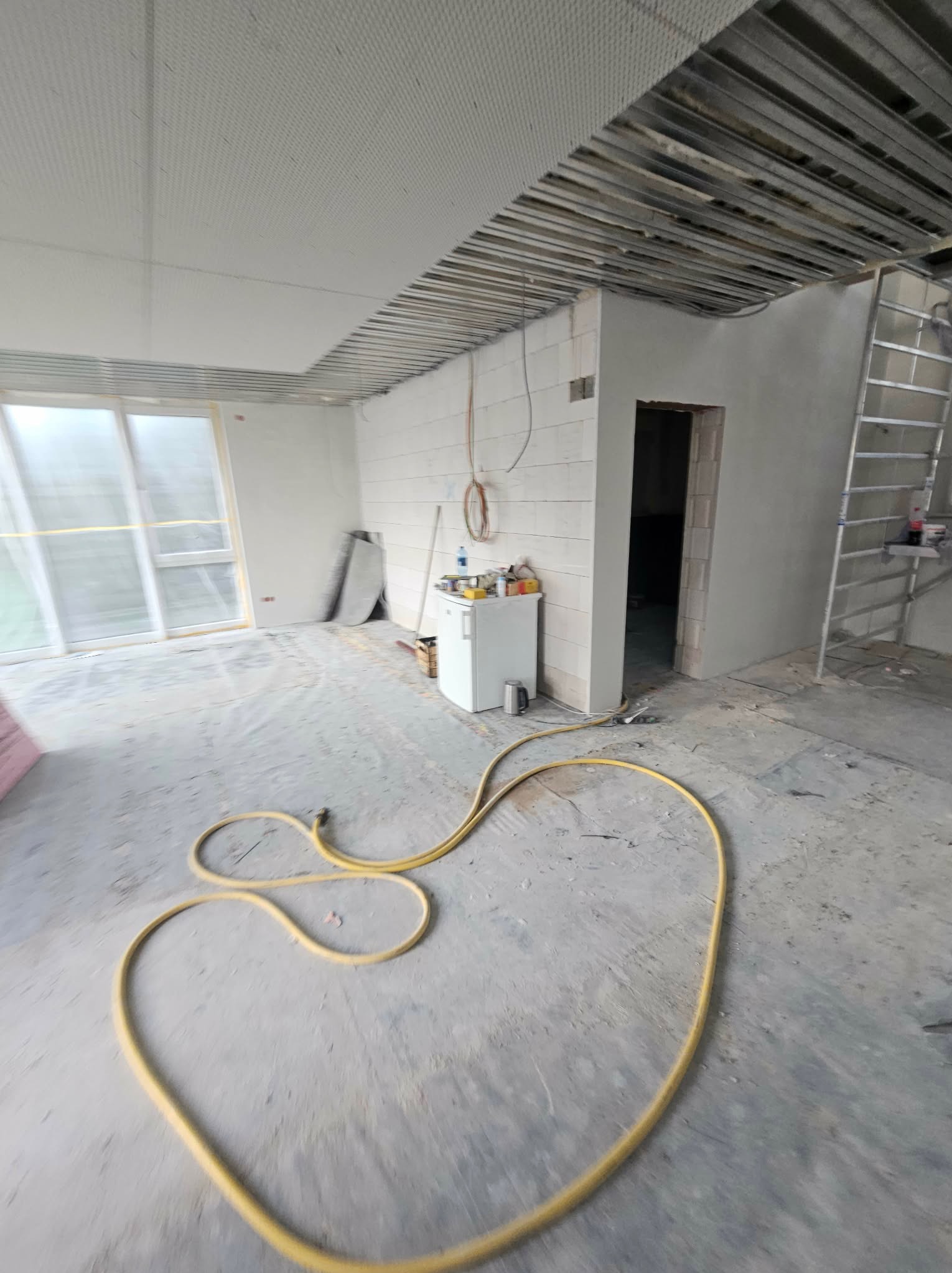 Wnętrze w trakcie remontu: widoczne profile sufitowe, ściany z bloków, okno, drabina i żółty wąż na podłodze. Etap prac wykończeniowych w pomieszczeniu.