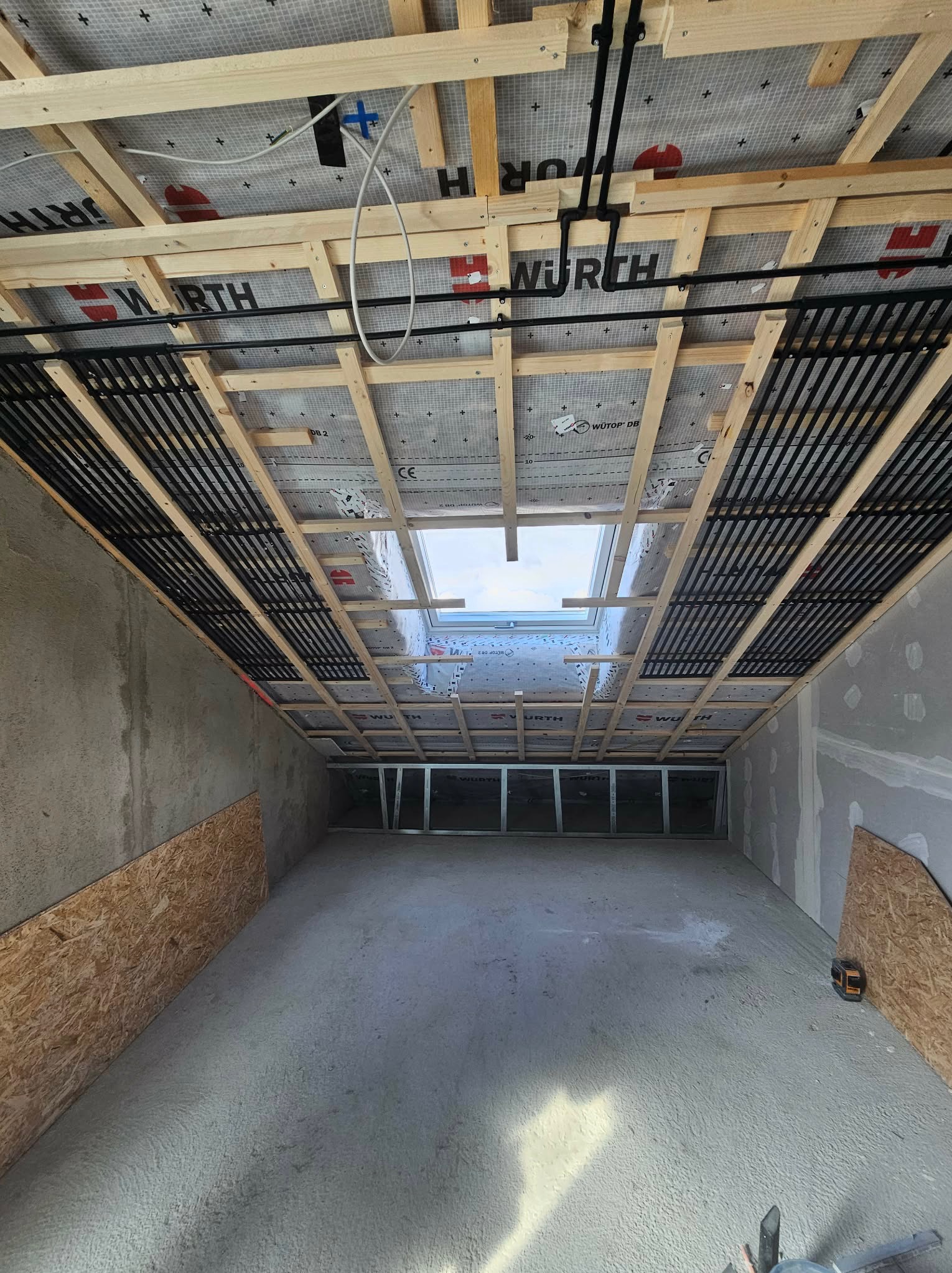 Montaż stelażu pod płytę gipsową na poddaszu z oknem dachowym. Widoczne drewniane belki, czarne rury instalacyjne i fragment ściany z płytą OSB.