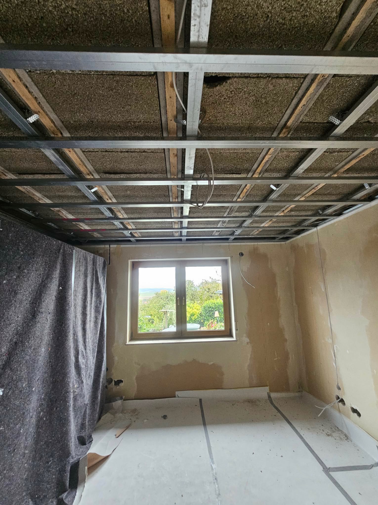 Montaż sufitu podwieszanego: metalowa konstrukcja z izolacją, widok z dołu na okno i przygotowane ściany. Etap prac wykończeniowych w jasnym pomieszczeniu.