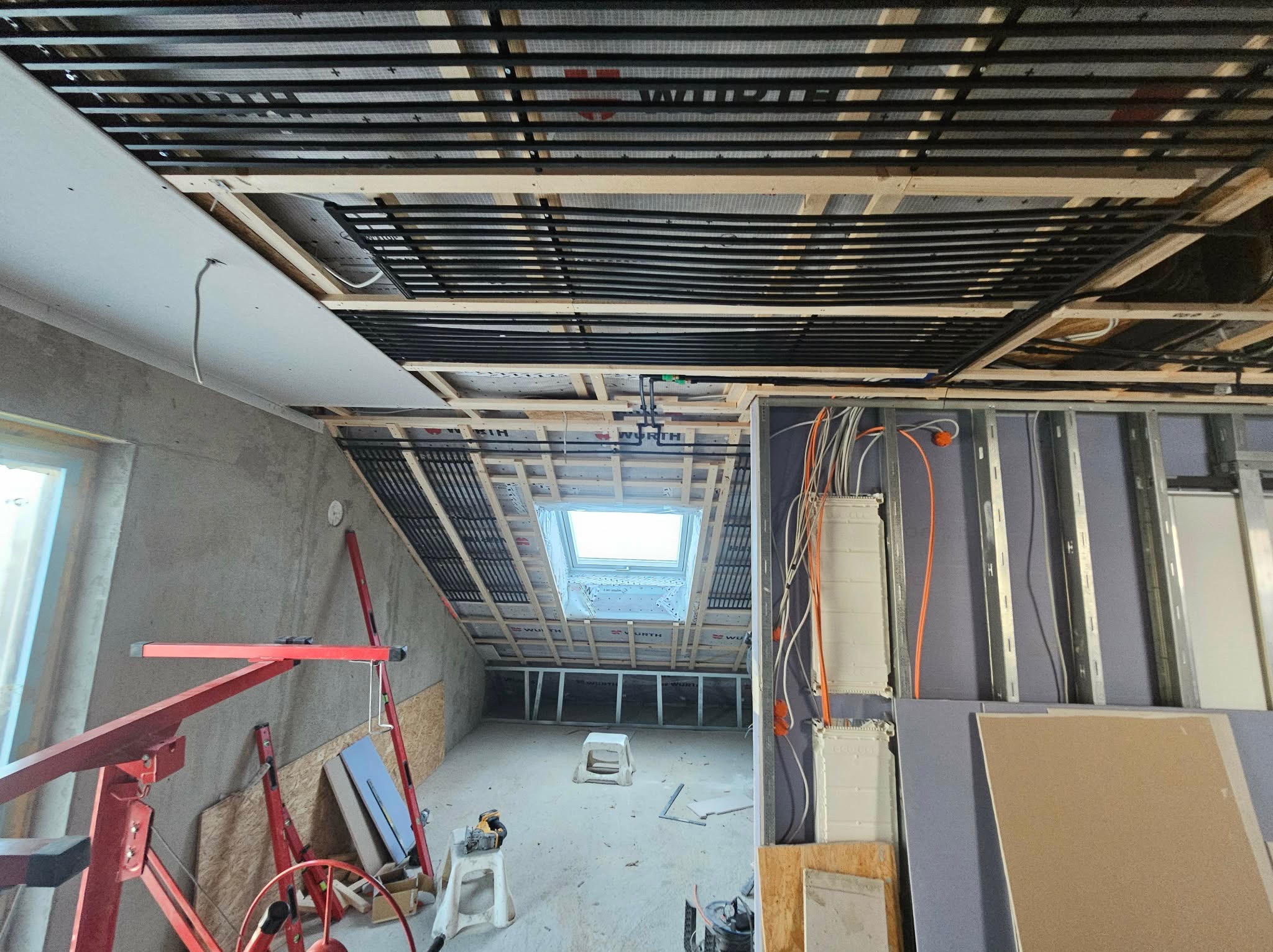 Montaż ogrzewania poddasza z widoczną instalacją pod płytami gipsowymi i oknem dachowym. Widoczne narzędzia i elementy konstrukcyjne.