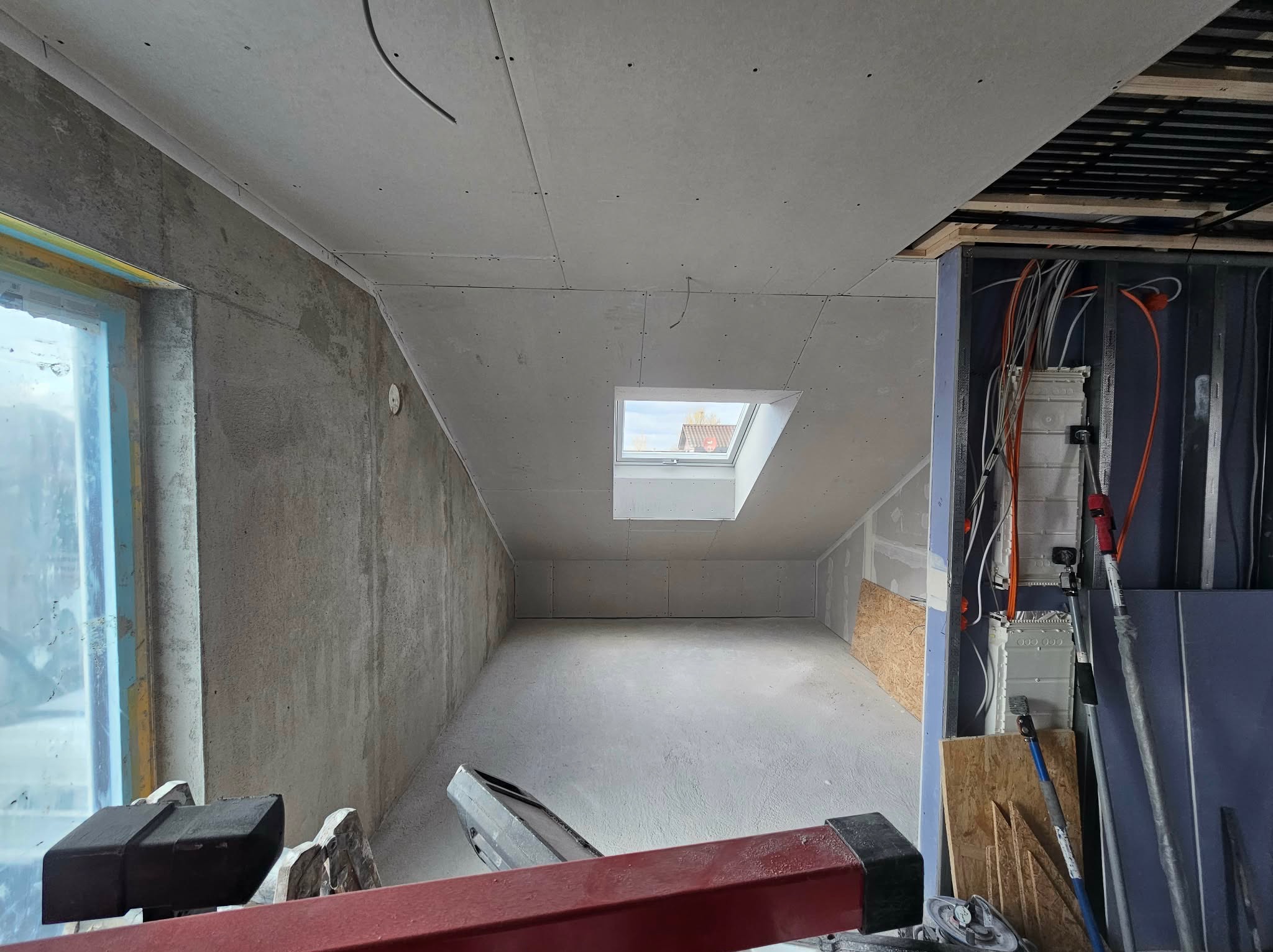 Surowe wnętrze poddasza z oknem dachowym i ścianami wykończonymi płytami gipsowo-kartonowymi. Widoczne instalacje elektryczne i fragmenty konstrukcji nośnej.