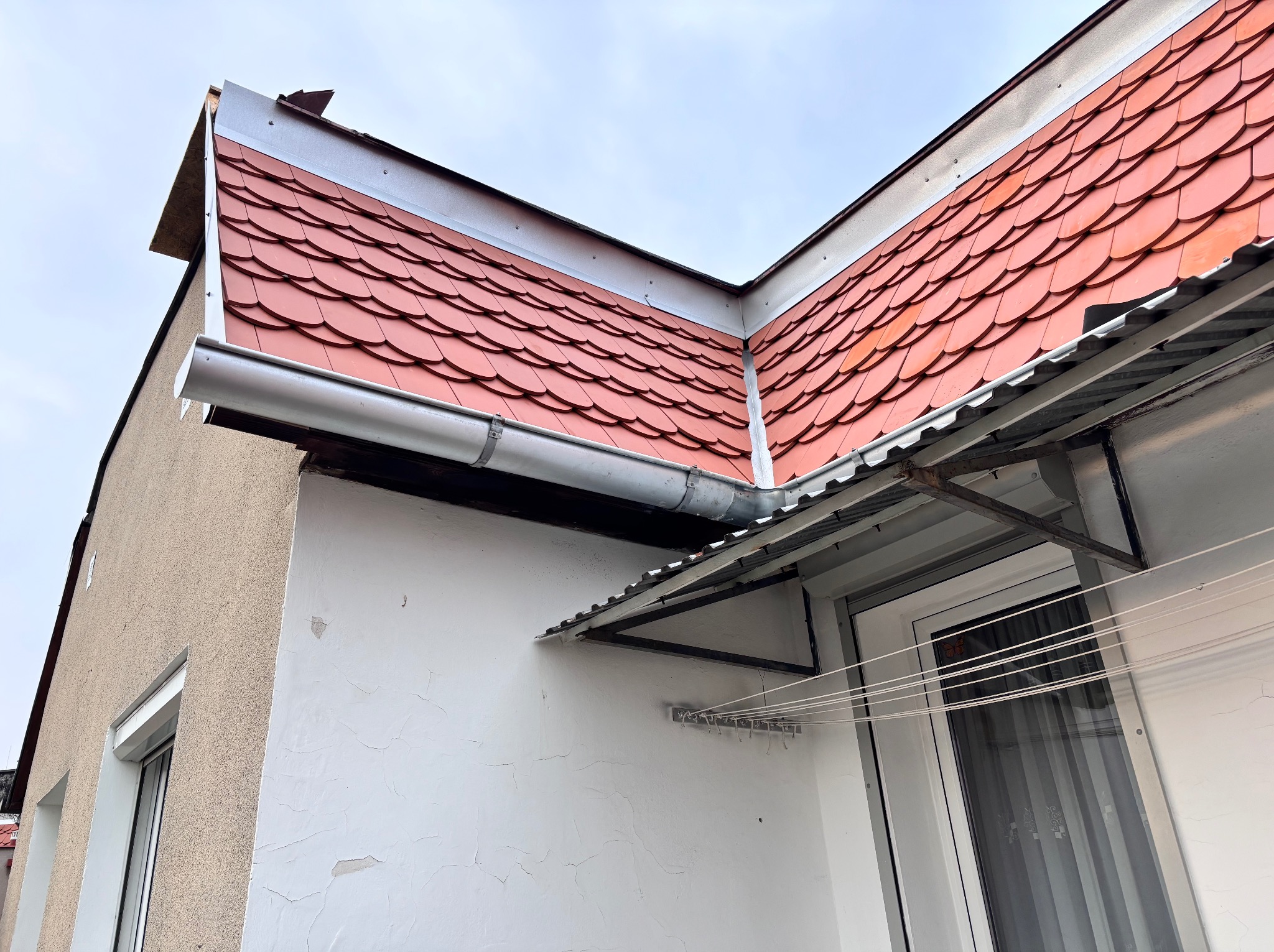 Czerwona dachówka karpiówka na dachu budynku z zamontowaną rynną. Widoczna elewacja budynku z oknem i suszarką na pranie.