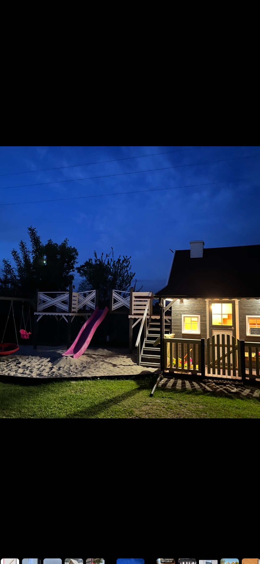 Oświetlony plac zabaw z domkiem, zjeżdżalnią i huśtawką, otoczony piaskiem i trawnikiem, sfotografowany o zmierzchu. Widoczne detale konstrukcji i oświetlenia.