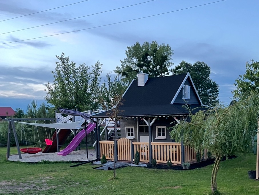 Drewniany domek zabaw dla dzieci z huśtawką i zjeżdżalnią, otoczony zielenią. Domek pomalowany na szaro z białymi detalami, czarny dach. Widok z poziomu trawnika.