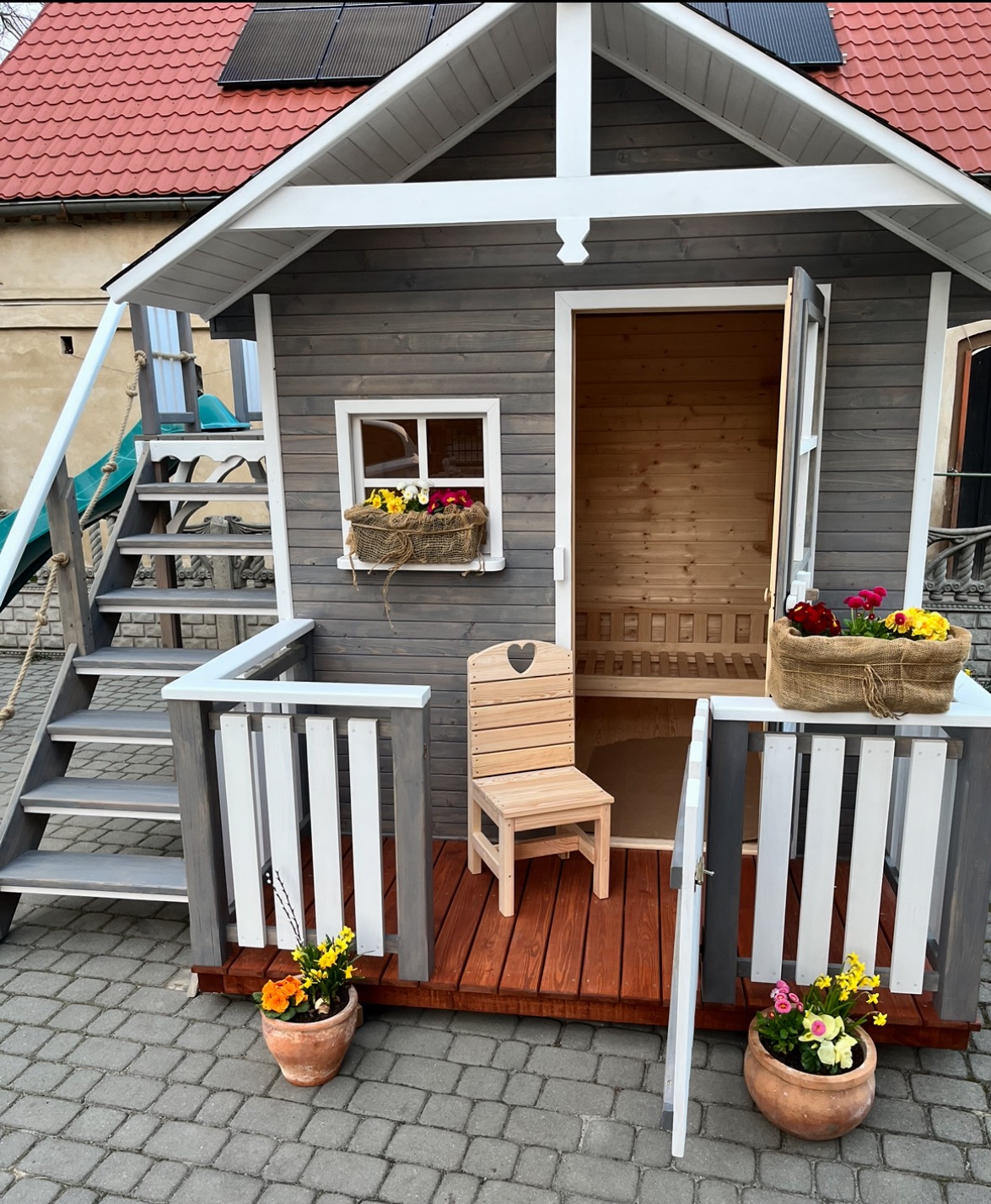 Drewniany domek dla dzieci z schodami, zjeżdżalnią i małym tarasem. Szare ściany, czerwony dach, kwiaty w doniczkach. Krzesło z sercem na werandzie.