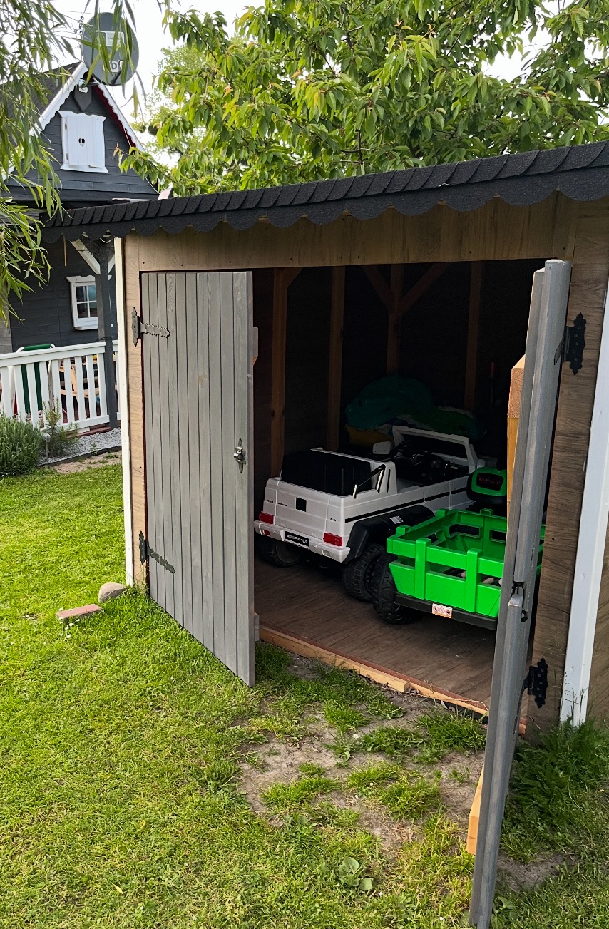 Drewniany domek narzędziowy z otwartymi drzwiami, wewnątrz zabawki dziecięce: biały Mercedes i zielony traktor. W tle dom z białym płotem i zielony trawnik.