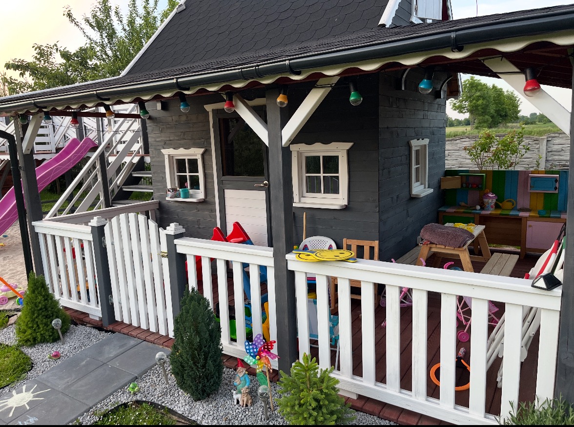 Szara drewniana chatka dla dzieci z białym ogrodzeniem i kolorowymi lampkami. W tle widoczna zjeżdżalnia i inne elementy placu zabaw. Ujęcie z bliska, w słoneczny dzień.
