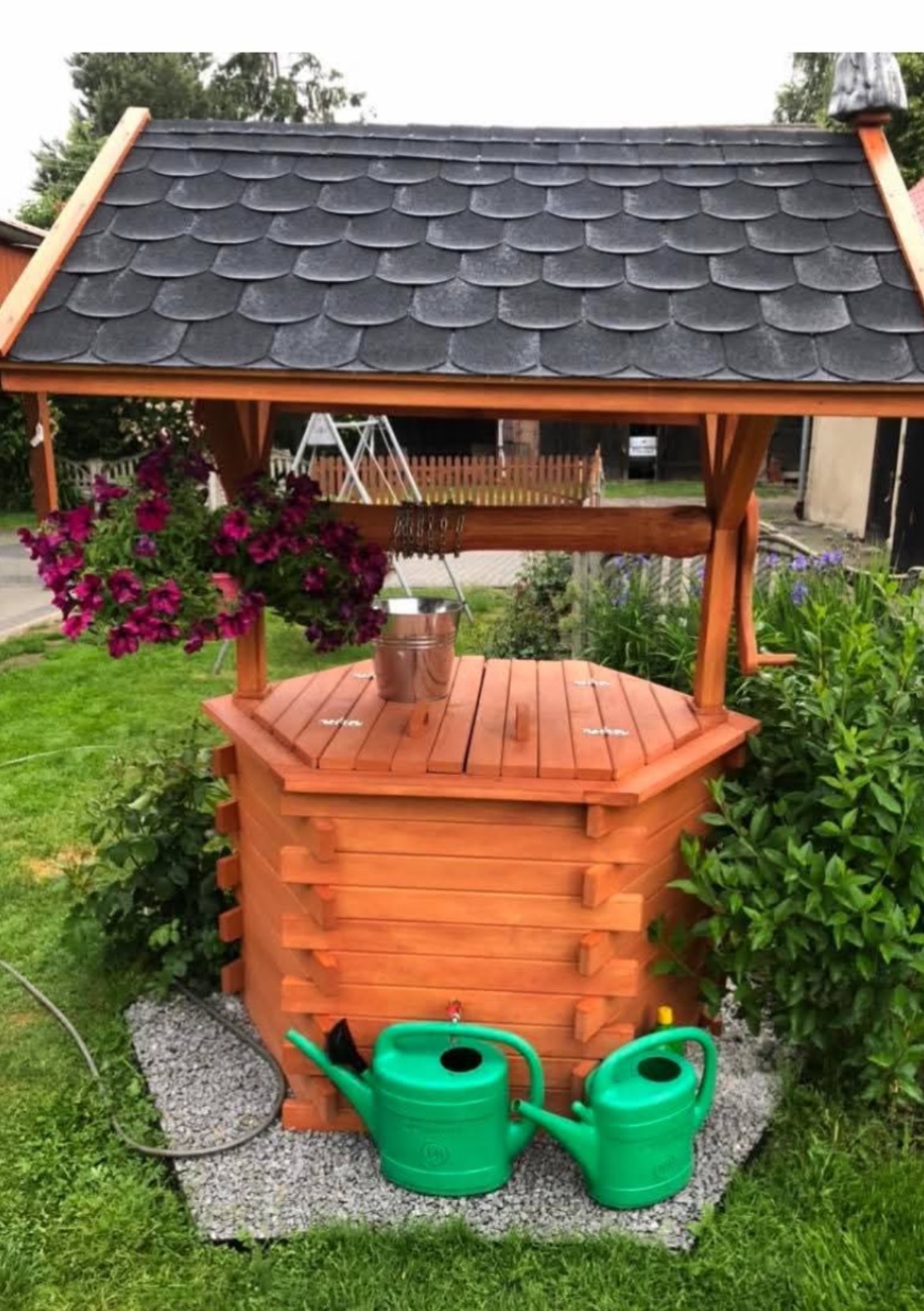 Drewniana studnia ogrodowa z czarnym dachem krytym gontem, ozdobiona kwiatami i zielonymi konewkami, otoczona zielenią. W tle huśtawka i ogrodzenie.