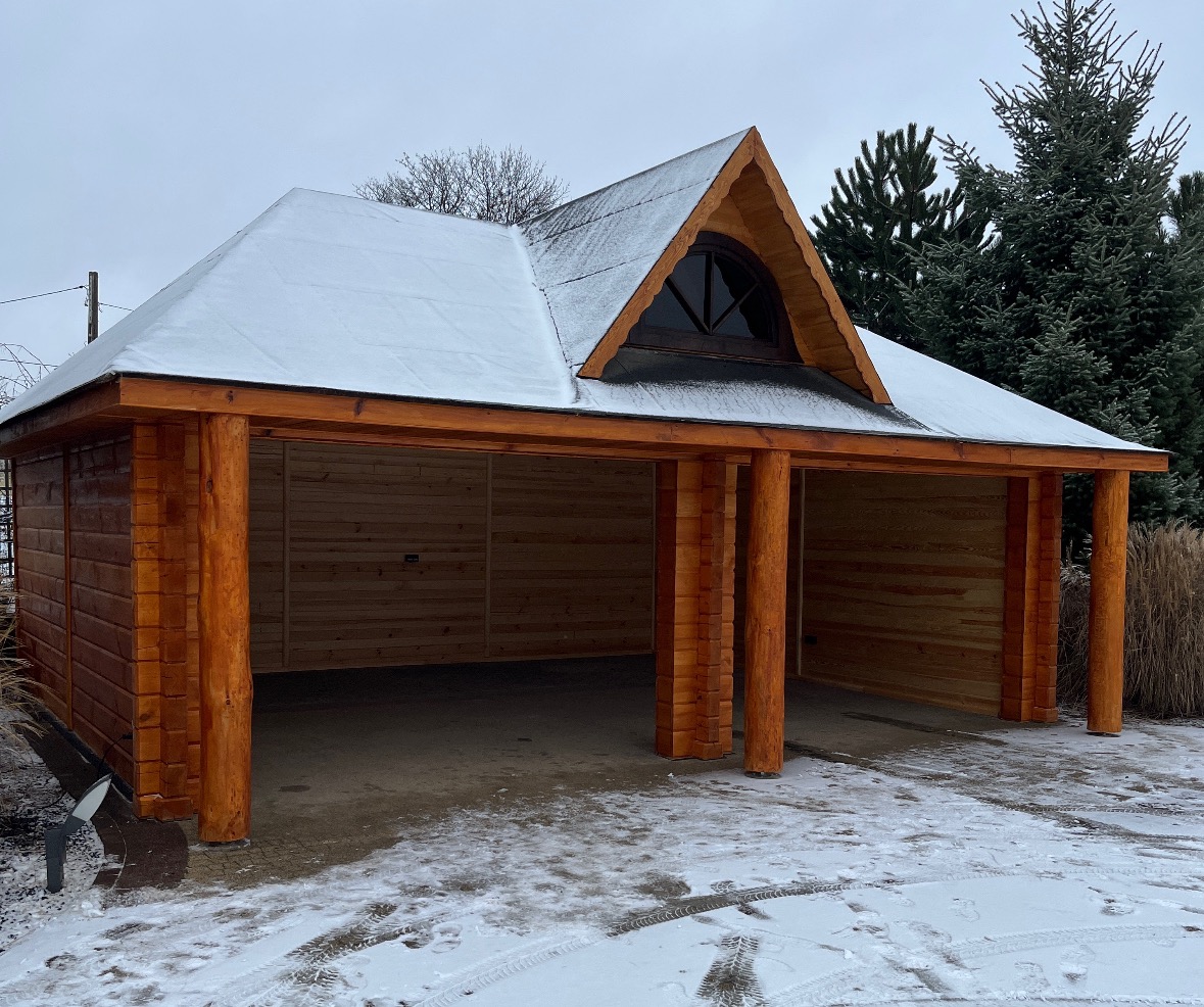 Drewniana wiata garażowa z dachem pokrytym śniegiem, widoczna z zewnątrz w zimowej scenerii. Konstrukcja z belek, zadaszenie z małym oknem na szczycie.