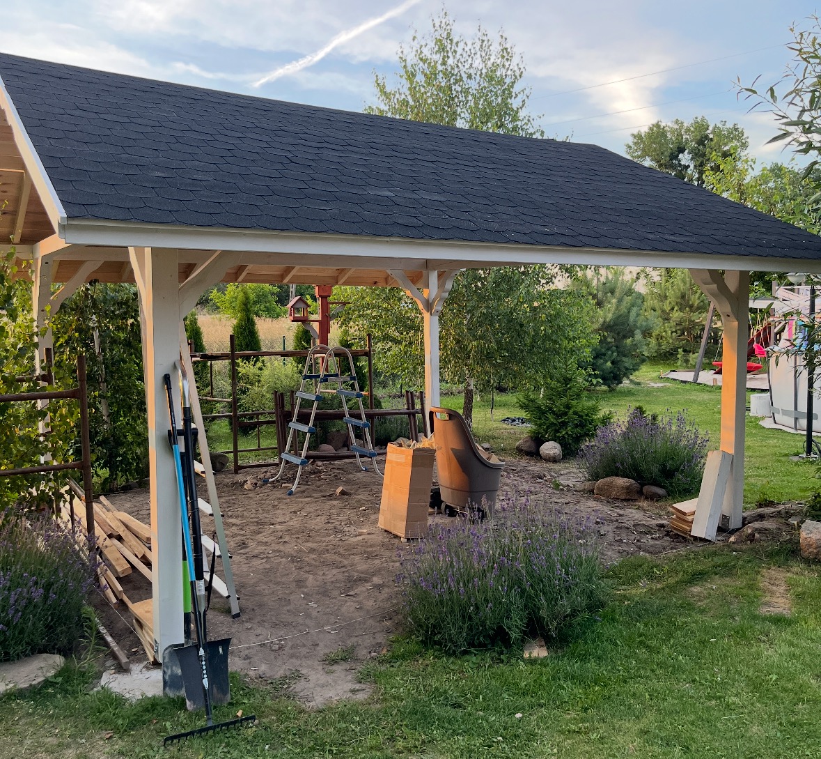 Zadaszona konstrukcja z czarnym dachem z gontu bitumicznego i białymi słupami, w trakcie budowy, z narzędziami i materiałami budowlanymi w tle ogrodu.