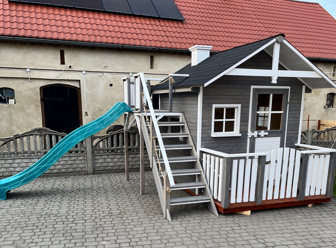 Drewniany domek zabaw dla dzieci z turkusową zjeżdżalnią, schodami i białym ogrodzeniem, na tle brukowanej nawierzchni i budynku z czerwoną dachówką.