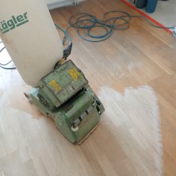 PARKIETEX KRZYSZTOF GRZEGORCZYK - Parkiet w trakcie renowacji z użyciem cykliniarki Lägler. Widoczny fragment podłogi po obróbce, kabel zasilający i akcesoria w tle. Jasne wnętrze.