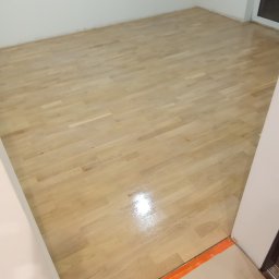 PARKIETEX KRZYSZTOF GRZEGORCZYK - Jasny, lakierowany parkiet w pustym pokoju z białymi ścianami. Widoczny fragment okna i pomarańczowe zabezpieczenie przy progu. Podłoga po renowacji, gotowa do użytku.