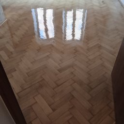 PARKIETEX KRZYSZTOF GRZEGORCZYK - Parkiet ułożony w jodełkę, po renowacji. Widoczne odbicie okna w lakierowanej powierzchni. Jasne drewno, świeżo po nałożeniu lakieru. Widok z progu.