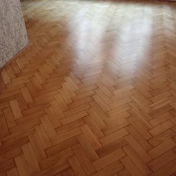 PARKIETEX KRZYSZTOF GRZEGORCZYK - Parkiet ułożony w jodełkę, po renowacji. Widoczne naturalne usłojenie drewna i połysk lakieru. W tle ściana z tapetą i grzejnik.