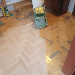 Cyklinowanie podłóg - Parkiet w jodełkę częściowo odnowiony, z widoczną maszyną do cyklinowania i odkurzaczem przemysłowym. Kontrast między starą a nową powierzchnią.