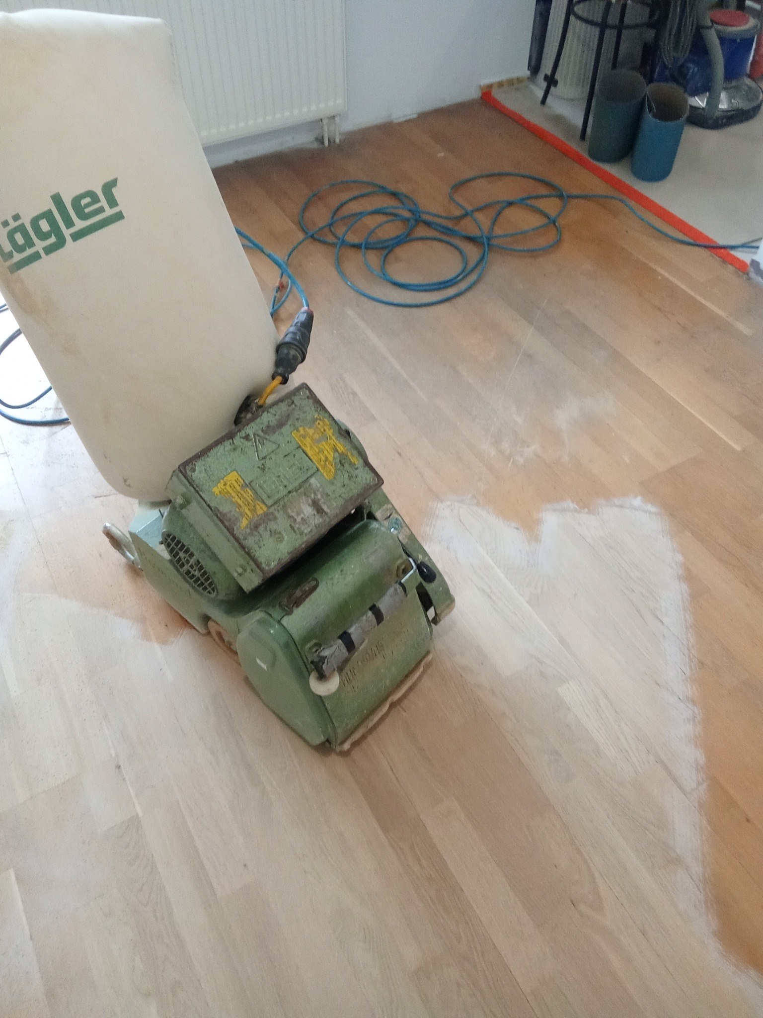 Parkiet w trakcie renowacji z użyciem cykliniarki Lägler. Widoczny fragment podłogi po obróbce, kabel zasilający i akcesoria w tle. Jasne wnętrze.