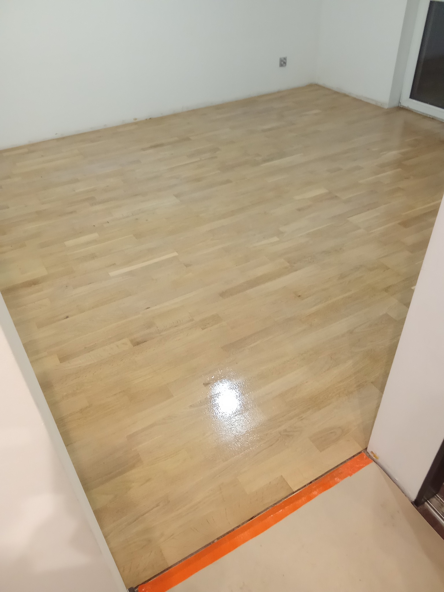 Jasny, lakierowany parkiet w pustym pokoju z białymi ścianami. Widoczny fragment okna i pomarańczowe zabezpieczenie przy progu. Podłoga po renowacji, gotowa do użytku.
