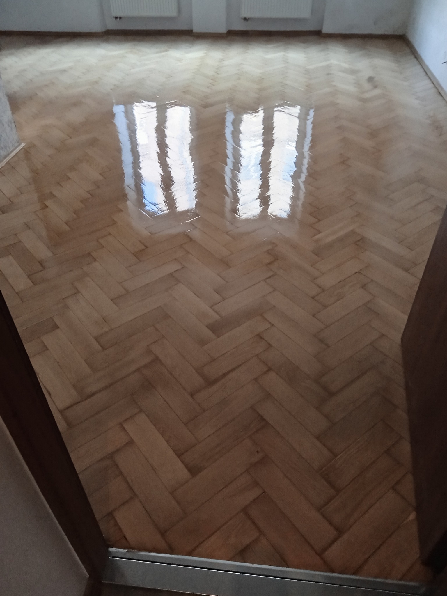 Parkiet ułożony w jodełkę, po renowacji. Widoczne odbicie okna w lakierowanej powierzchni. Jasne drewno, świeżo po nałożeniu lakieru. Widok z progu.