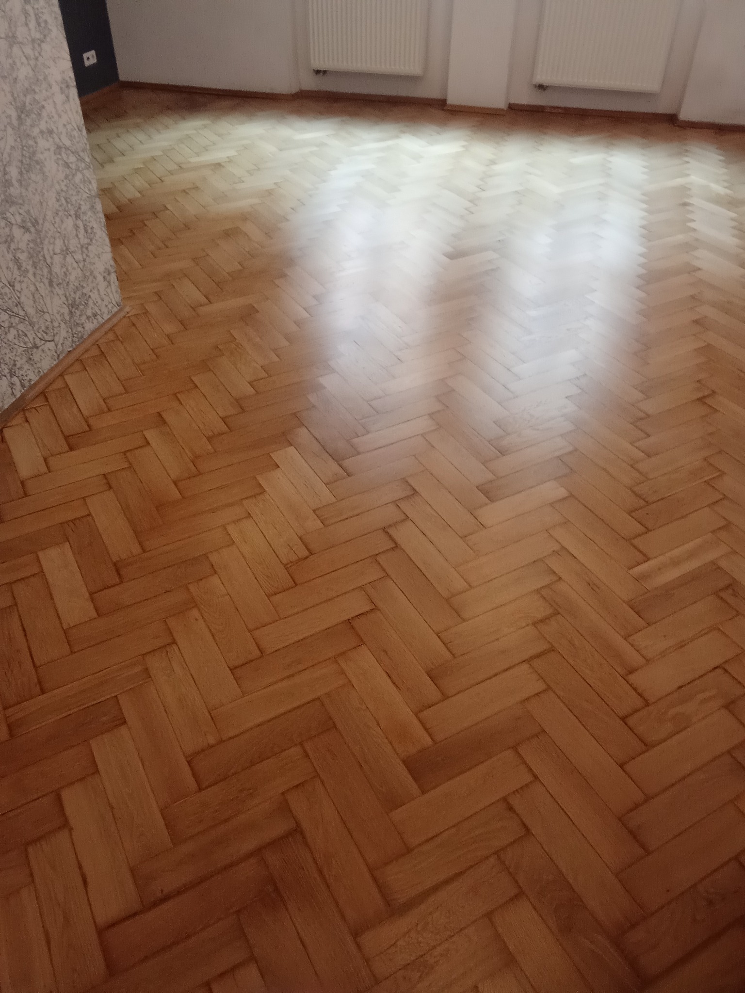 Parkiet ułożony w jodełkę, po renowacji. Widoczne naturalne usłojenie drewna i połysk lakieru. W tle ściana z tapetą i grzejnik.