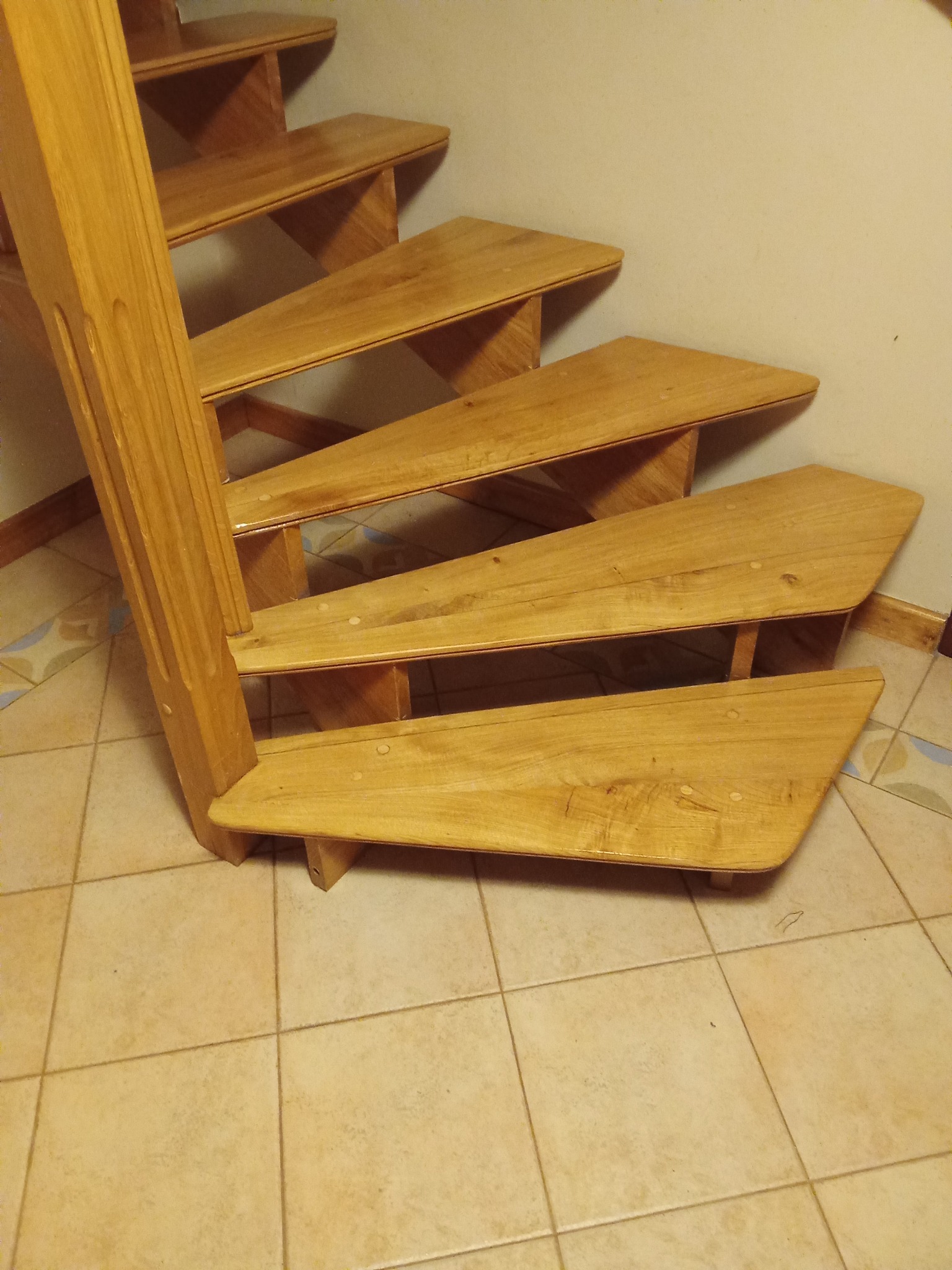 Drewniane schody z jasnego drewna, widok z dołu, ukazujący stopnie i fragment poręczy. Na podłodze jasne płytki. Widoczne ślady po cyklinowaniu.