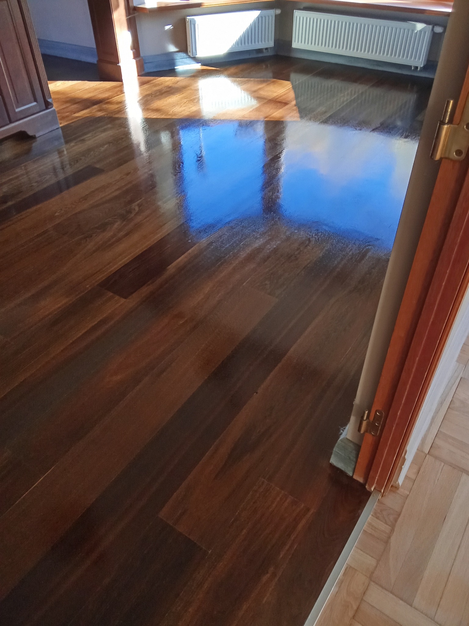 Błyszcząca, ciemna podłoga z parkietu odbija niebo, widoczna przez otwarte drzwi. Fragment grzejnika i okna w tle. Ujęcie z niskiego kąta.