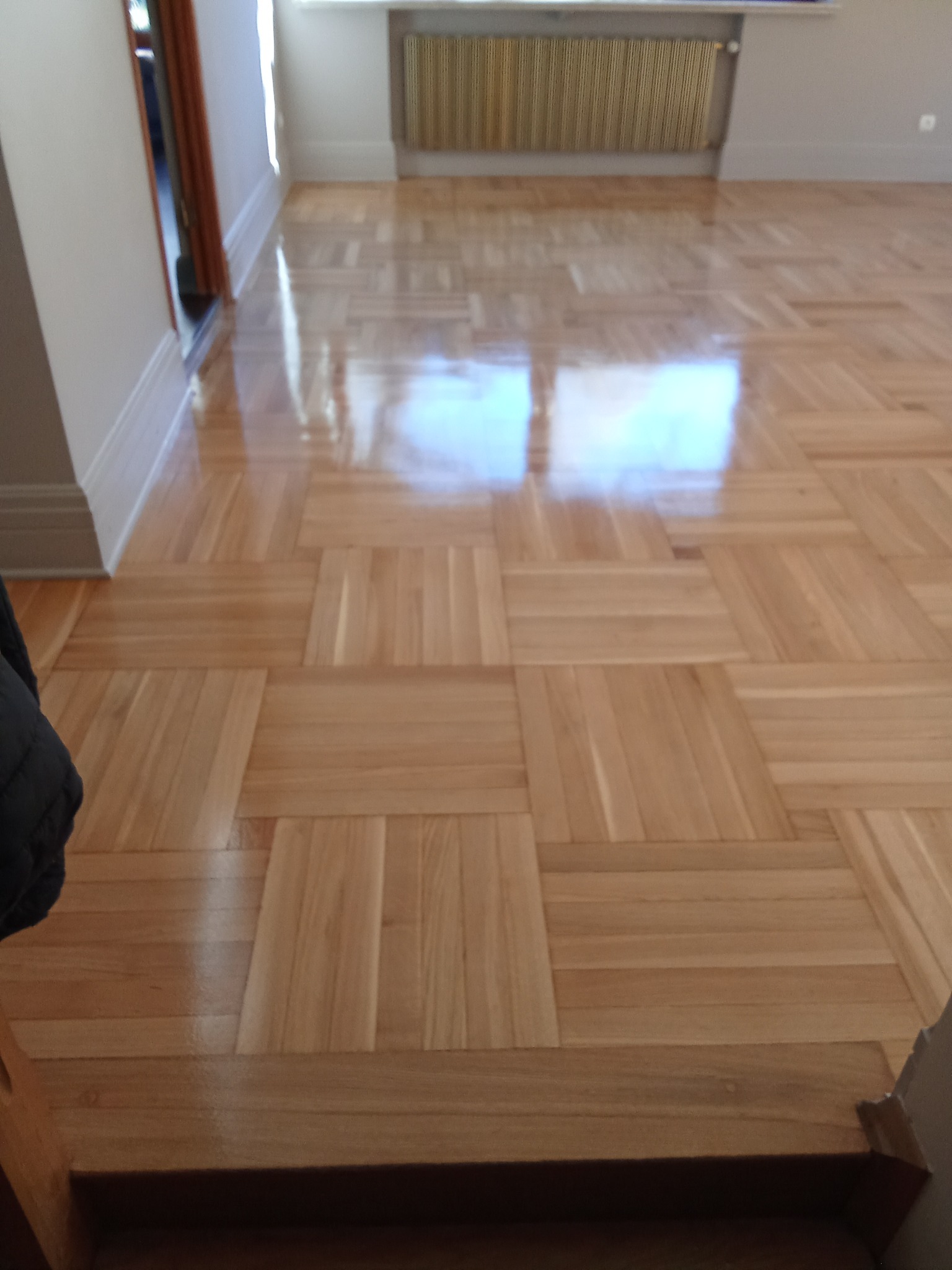 Błyszcząca, świeżo polakierowana podłoga parkietowa ułożona we wzór kwadratów, odbijająca światło okna. Widoczny fragment grzejnika i drzwi.