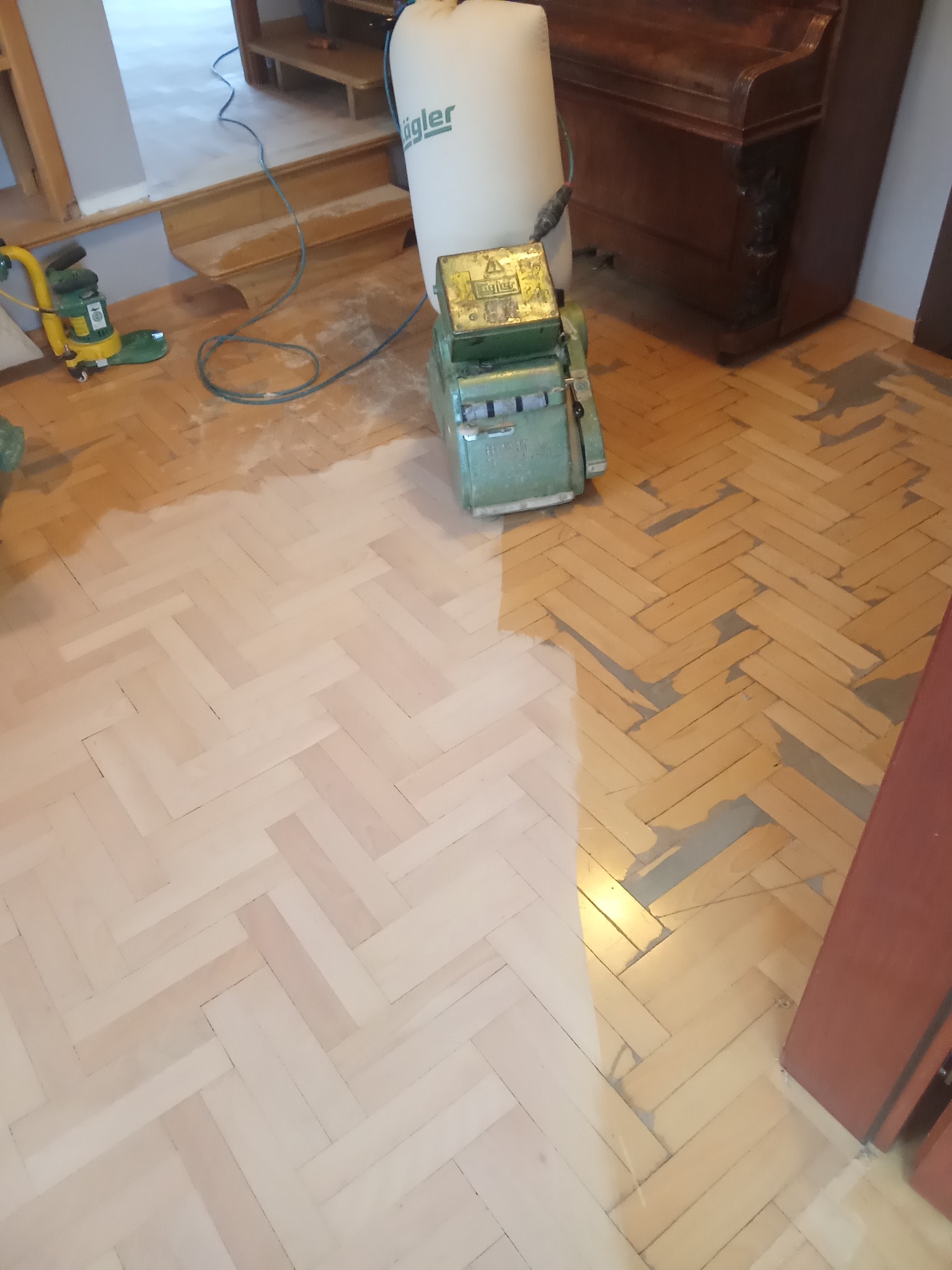 Parkiet w jodełkę częściowo odnowiony, z widoczną maszyną do cyklinowania i odkurzaczem przemysłowym. Kontrast między starą a nową powierzchnią.