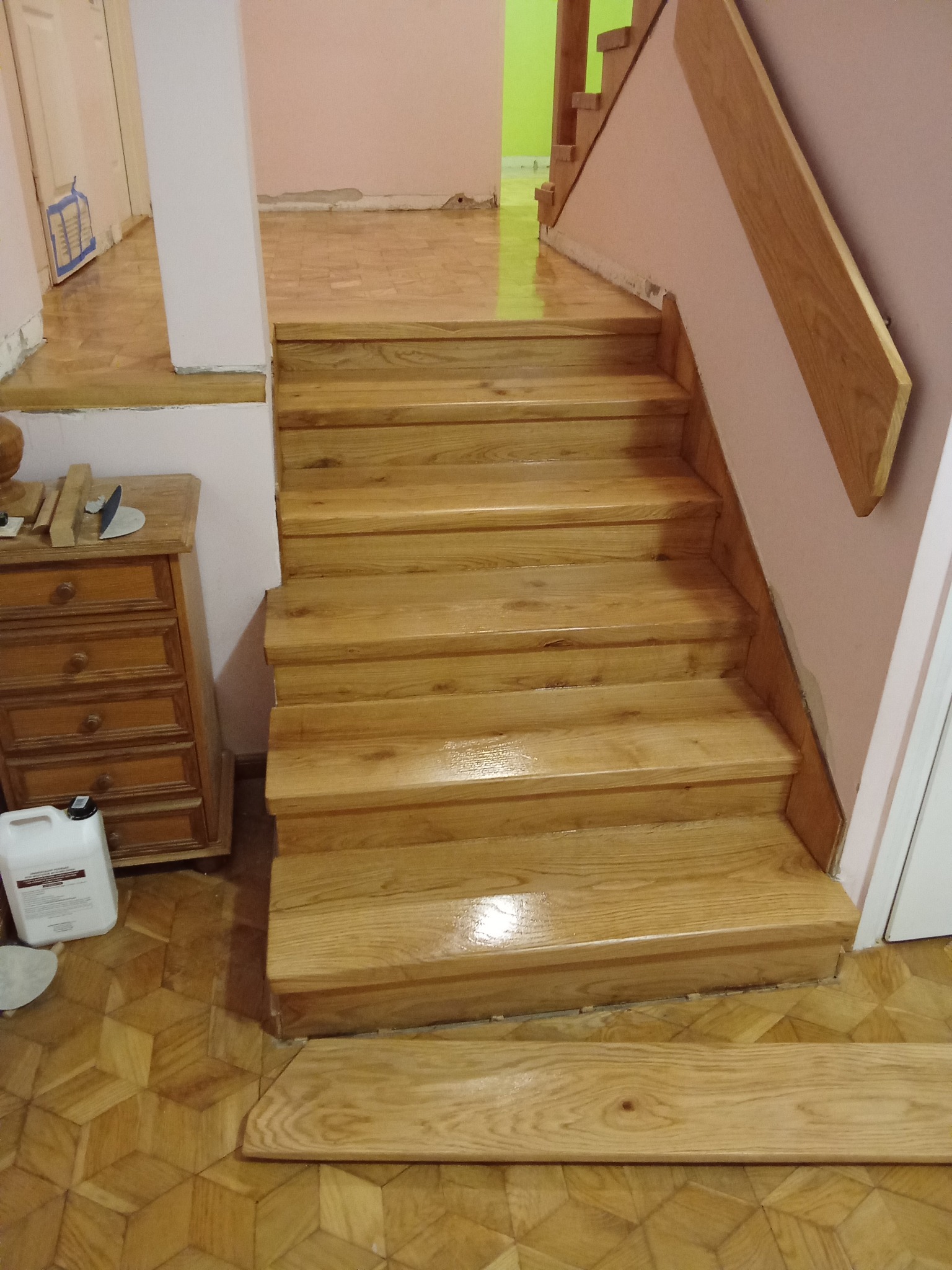 Odnowione drewniane schody z parkietem w jodełkę. Widoczna komoda z narzędziami i preparat do pielęgnacji drewna. Jasne, ciepłe oświetlenie.