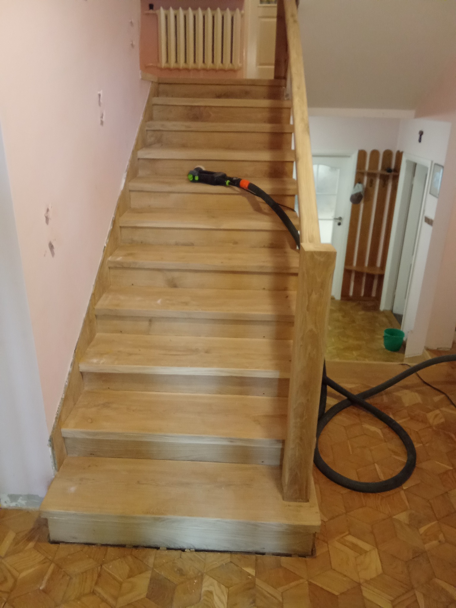 Jasne, drewniane schody po cyklinowaniu, z widocznym odkurzaczem przemysłowym i parkietem w jodełkę na podłodze. Ściany w odcieniach różu i beżu.