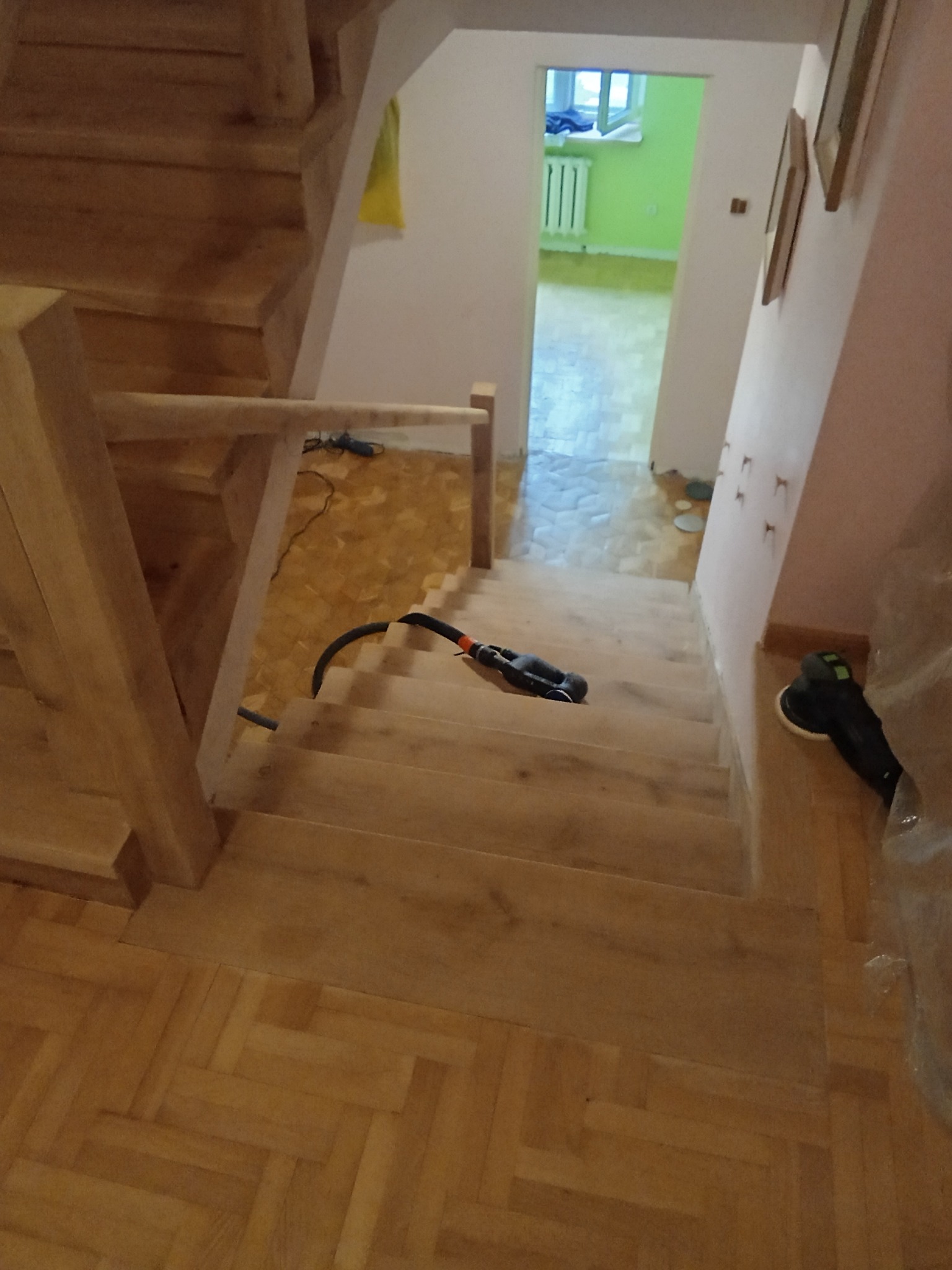 Drewniane schody w trakcie renowacji. Widoczny odkurzacz i szlifierka. Parkiet w jodełkę na podeście. Oświetlone wnętrze z zieloną ścianą w tle.