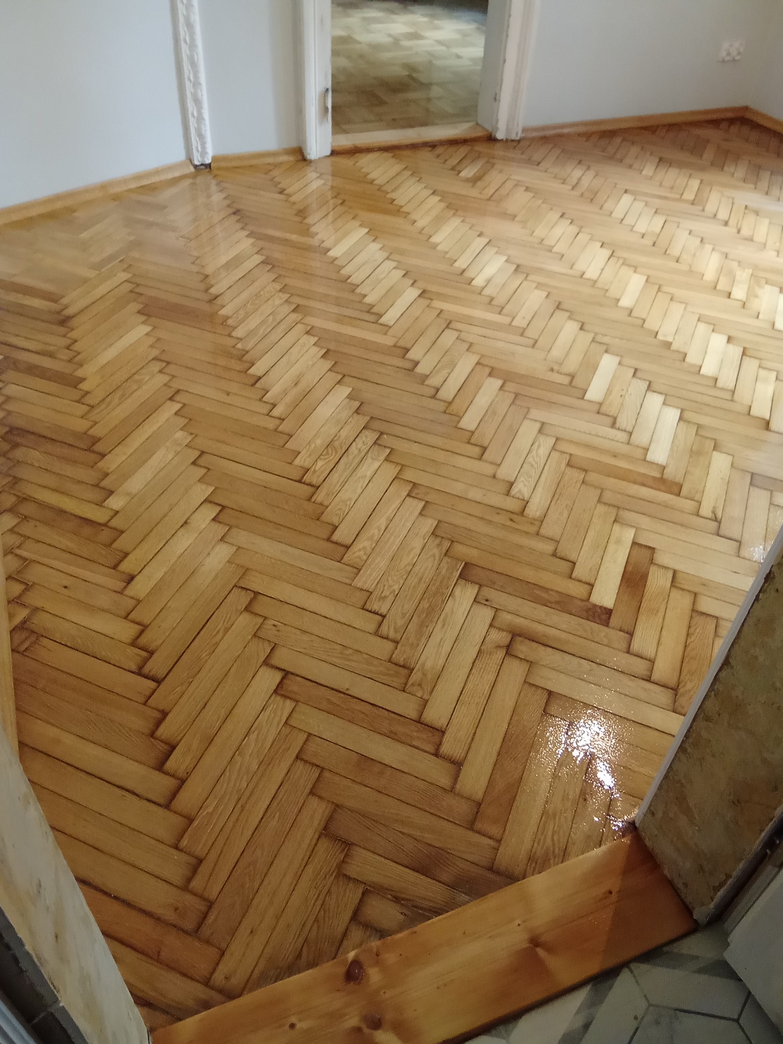 Parkiet ułożony w jodełkę, świeżo po lakierowaniu. Widoczne refleksy światła na gładkiej powierzchni. Jasne drewno, klasyczny wzór, wykończone wnętrze.