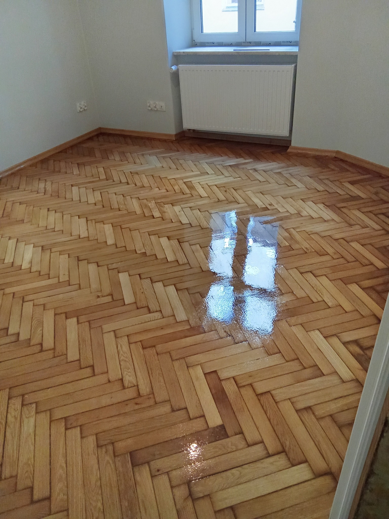 Parkiet ułożony w jodełkę, świeżo polakierowany, z widocznym odbiciem światła. Jasne drewno, listwy przypodłogowe, grzejnik pod oknem. Wykończenie podłogi.