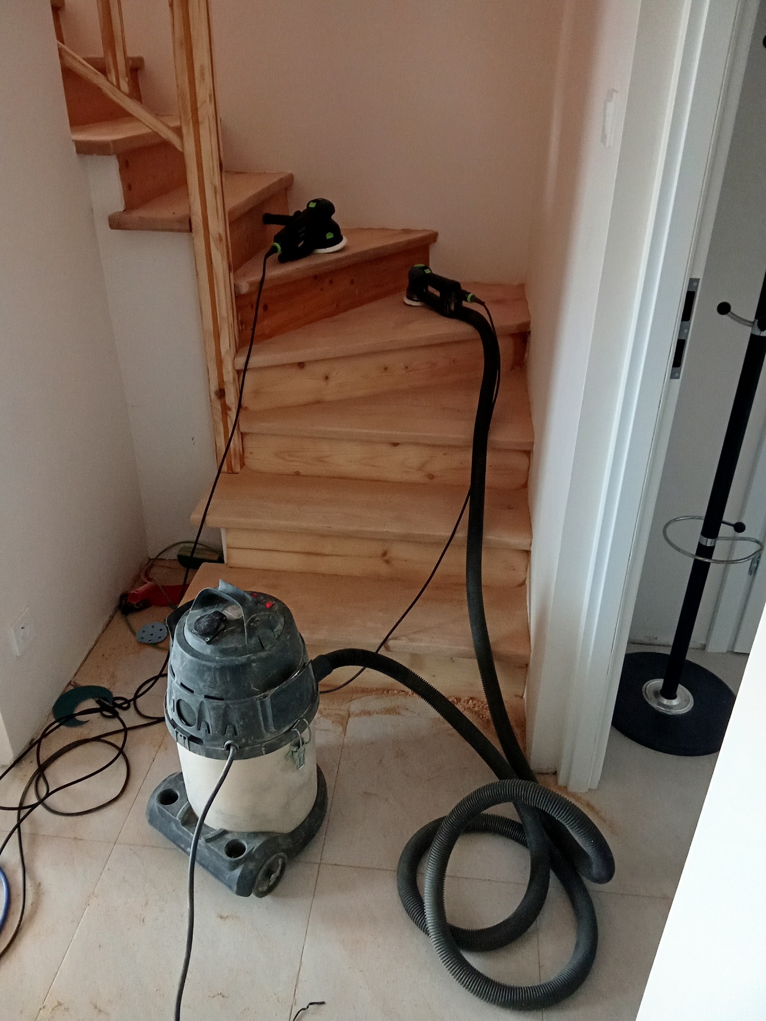 Drewniane schody w trakcie renowacji. Widoczny odkurzacz przemysłowy i szlifierka na schodach. Jasne drewno, białe ściany. Etap prac wykończeniowych.