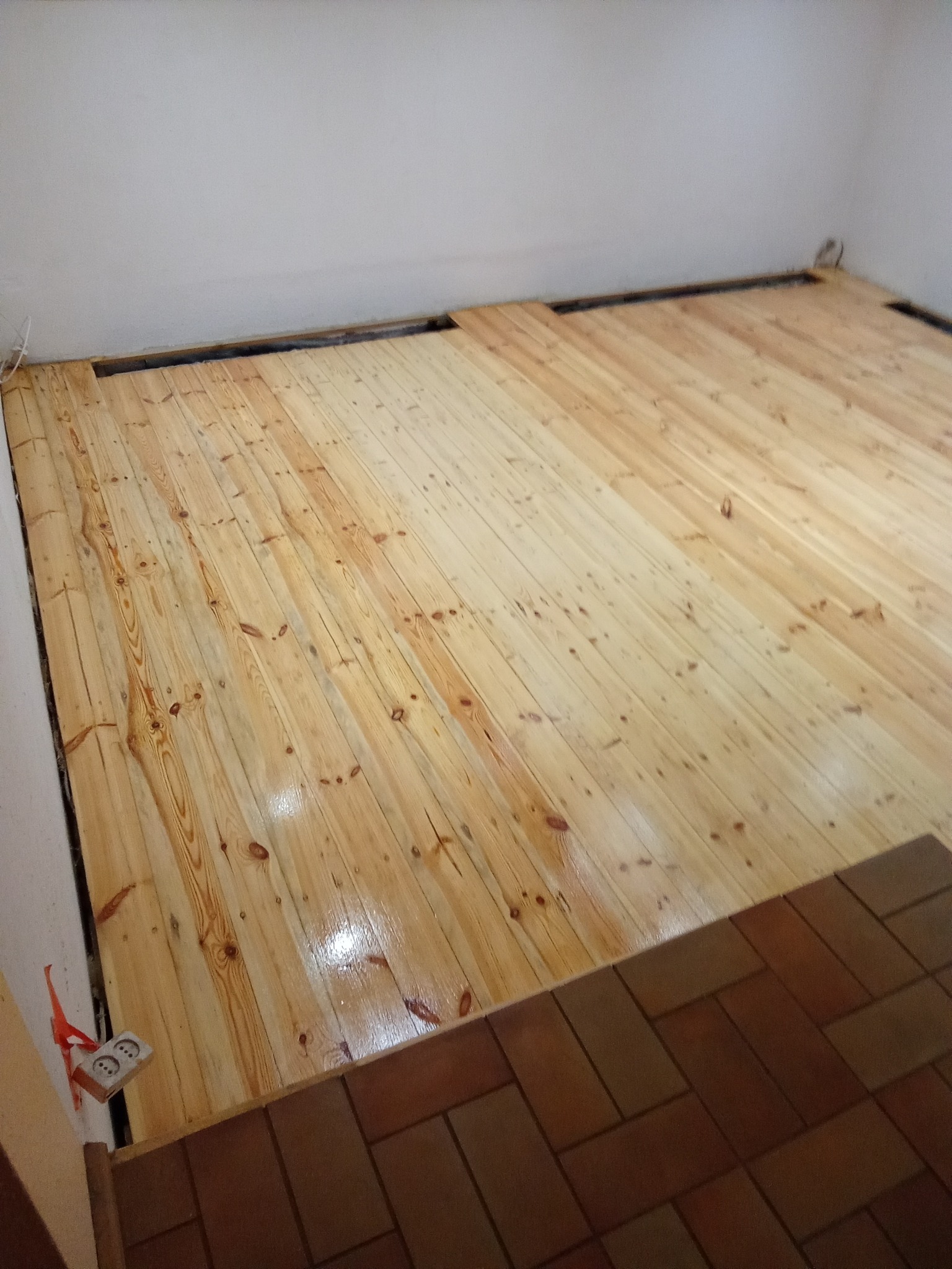 Lakierowana, jasna podłoga z desek w trakcie renowacji, widoczne szczeliny przy ścianach i gniazdko elektryczne. Część podłogi pokryta brązowymi płytkami.