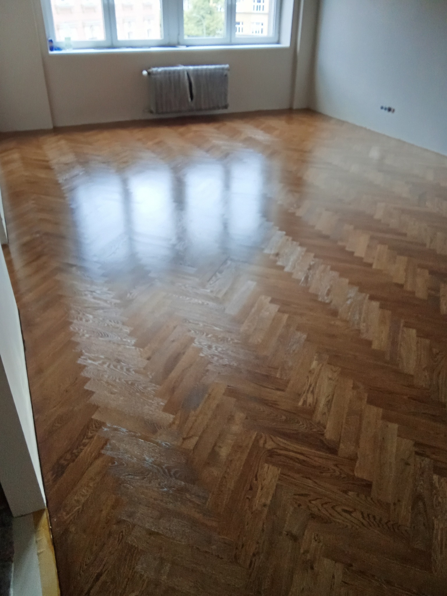 Parkiet ułożony w jodełkę w jasnym pokoju, widok z góry. Świeżo wykończona podłoga w mieszkaniu w Krakowie. Okno z grzejnikiem w tle.