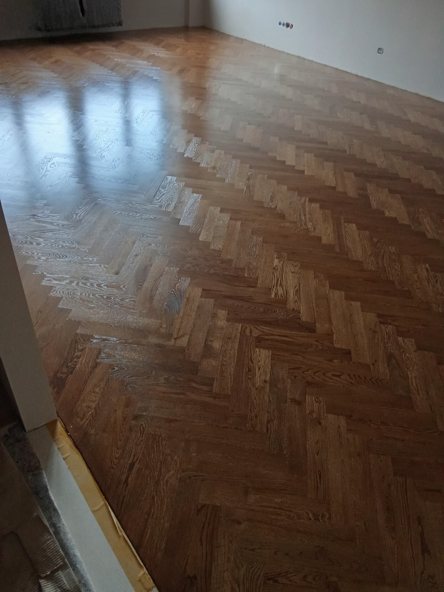 Ukończona podłoga z parkietu ułożonego w jodełkę, po renowacji. Widoczne refleksy światła na lakierowanej powierzchni. Detal wykończenia przy ścianie.