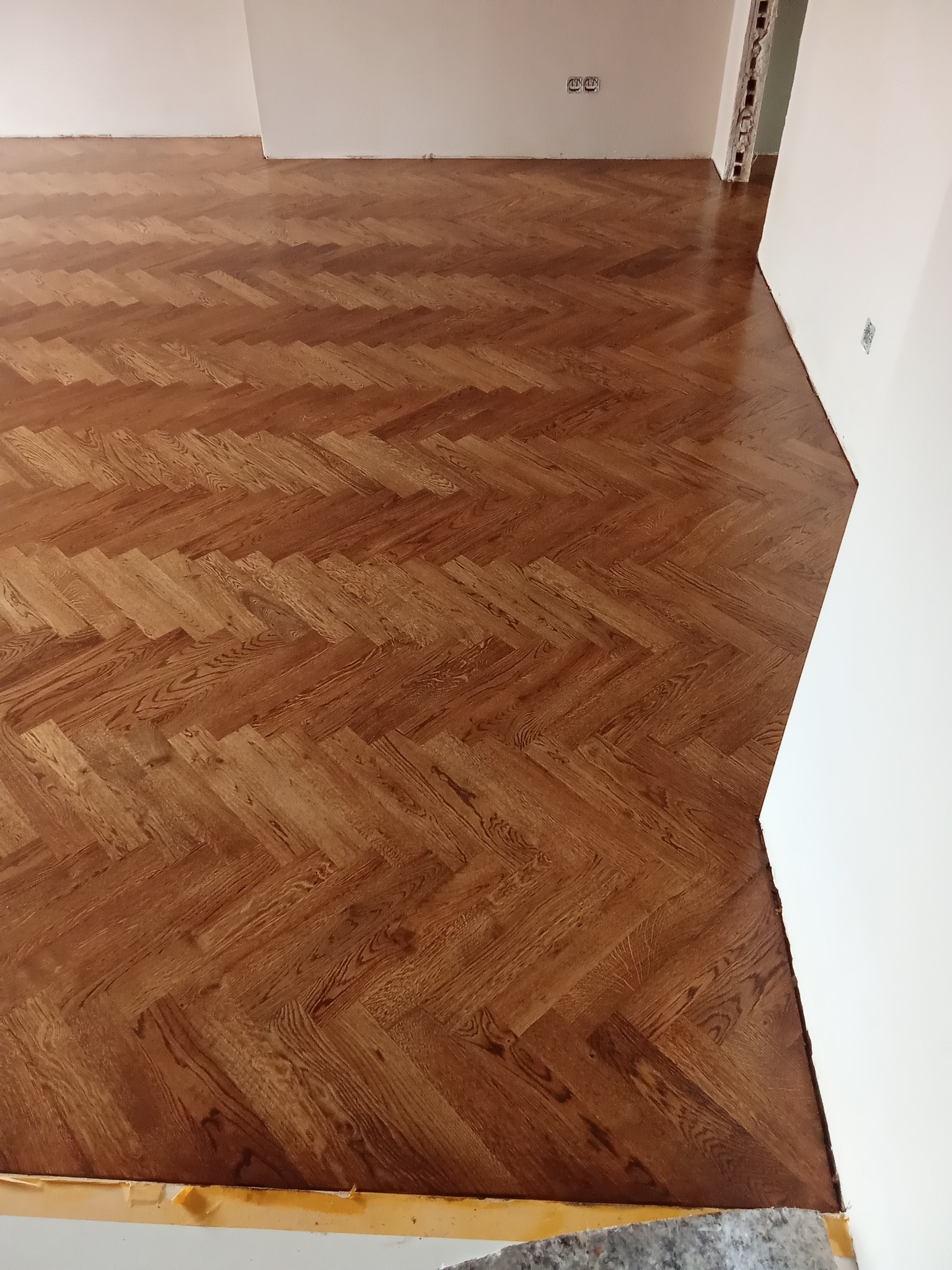 Ukończona podłoga z parkietu ułożonego w jodełkę, widok z góry, jasne ściany, fragment niezabezpieczonej podłogi przy ścianie.