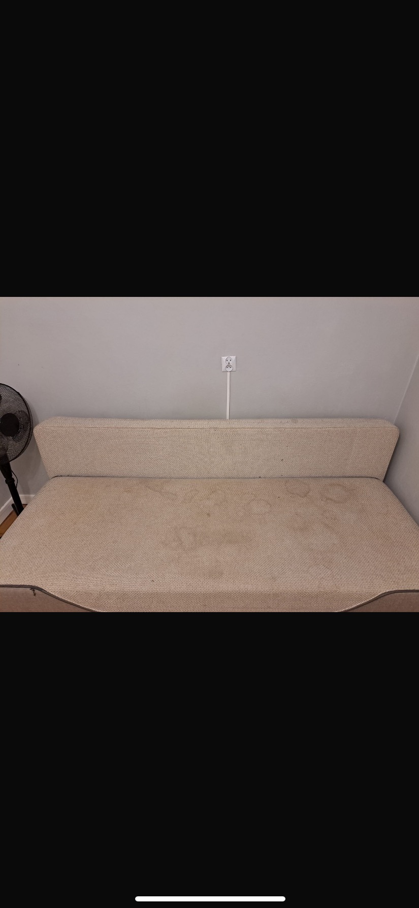 Beżowa kanapa z widocznymi plamami na tapicerce, stojąca na tle ściany z gniazdkiem elektrycznym i wentylatorem po lewej stronie. Widok z przodu, naturalne oświetlenie.