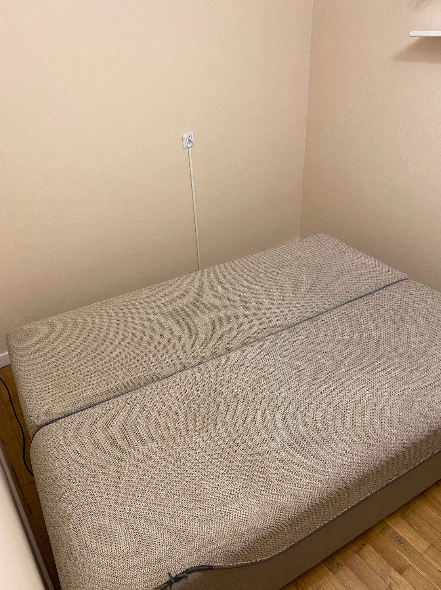 Rozłożona, beżowa kanapa z widoczną fakturą materiału tapicerskiego, ustawiona w rogu pokoju z drewnianą podłogą i gładkimi, beżowymi ścianami. Widoczne gniazdko elektryczne.