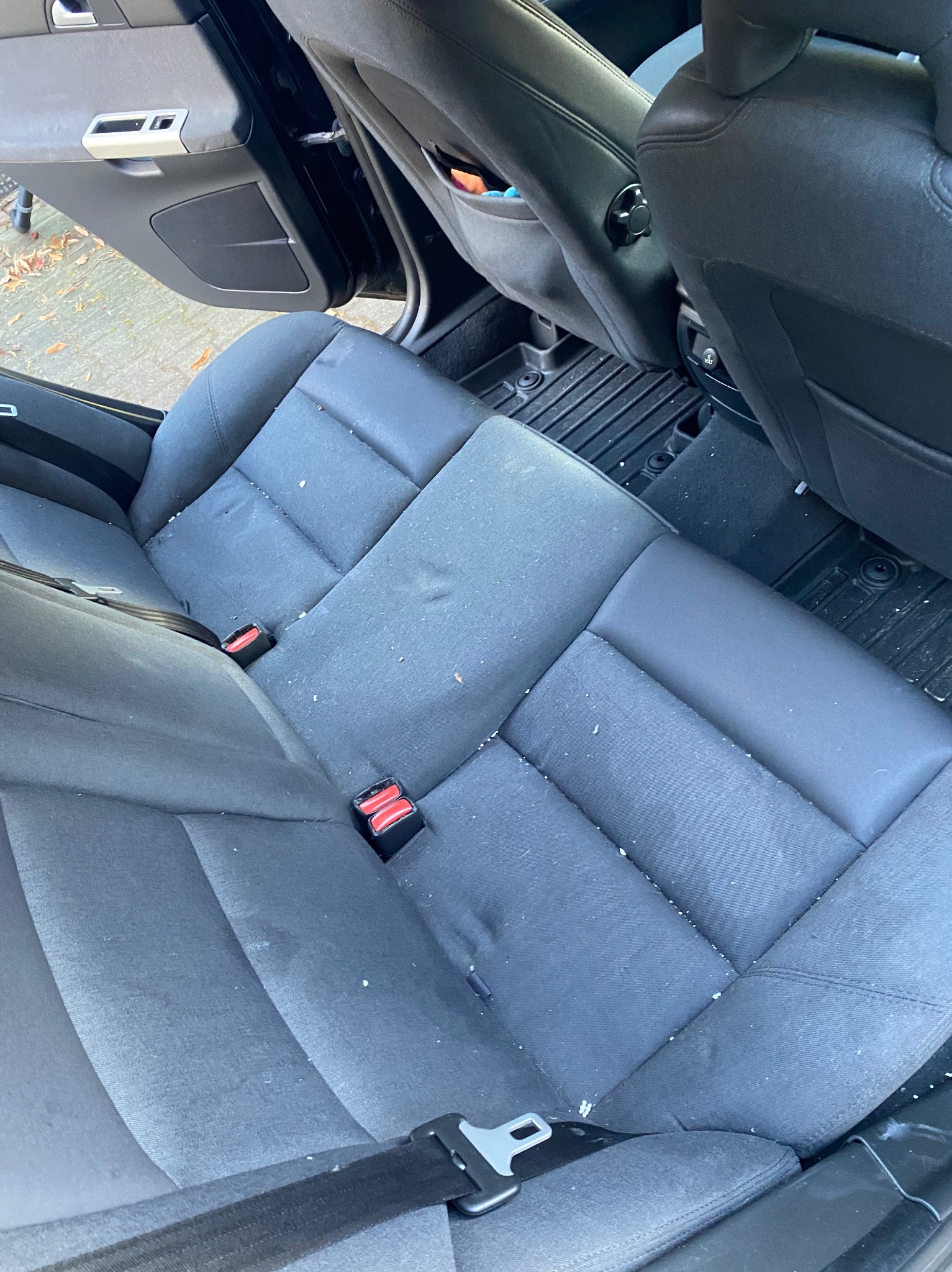 Szare, tekstylne siedzenie samochodowe z widocznymi plamami i zabrudzeniami, pasy bezpieczeństwa, czarna podłoga i fragment drzwi. Wnętrze pojazdu.