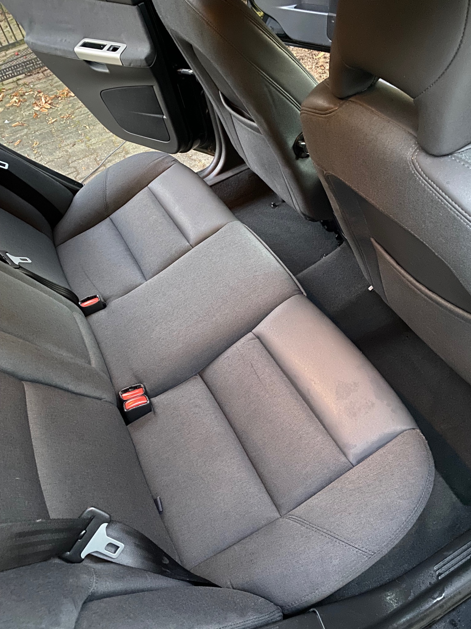 Szara, materiałowa tylna kanapa samochodowa z widocznymi pasami bezpieczeństwa i plamą na siedzisku, wnętrze pojazdu.