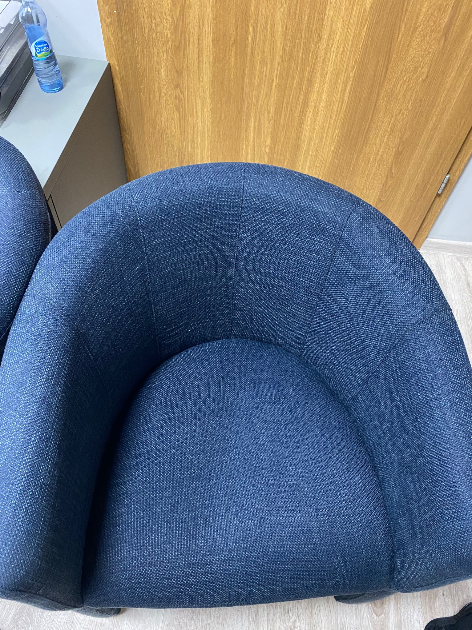Granatowe krzesło tapicerowane z bliska, widoczna faktura materiału. W tle fragment biura: butelka wody, szafka i drewniane drzwi. Ujęcie z góry.