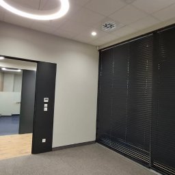 KS-BUD Serhii Koshil - Nowoczesne biuro po remoncie: szklane drzwi, czarne żaluzje, oświetlenie LED. Minimalistyczny design, jasne ściany, szara wykładzina. Widok na kolejne pomieszczenie.