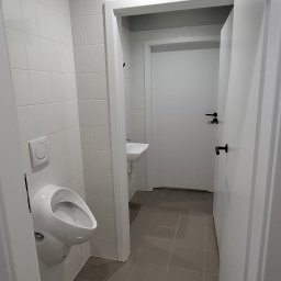 KS-BUD Serhii Koshil - Nowoczesna toaleta publiczna z białymi płytkami, wiszącym pisuarze i szarą podłogą z płytek. Widoczne drzwi i umywalka w tle. Minimalistyczny design.