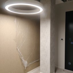 KS-BUD Serhii Koshil - Wnętrze w trakcie wykańczania: kolumna obłożona płytami, ściana zabezpieczona folią, okrągła lampa sufitowa. Widoczne gniazdka elektryczne i surowy sufit.