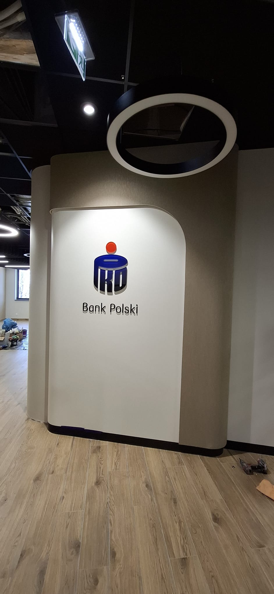 Wykończona ściana w biurze z logo IKO Bank Polski. Podłoga z paneli imitujących drewno. W tle widoczne okno i elementy wyposażenia biura. Nowoczesne oświetlenie sufitowe.
