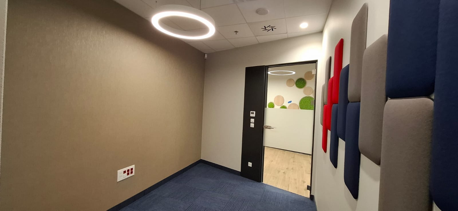 Nowoczesne wykończenie biura: beżowa ściana, niebieska wykładzina, drzwi z dekoracją roślinną i panele akustyczne w kolorach szarym, niebieskim i czerwonym.