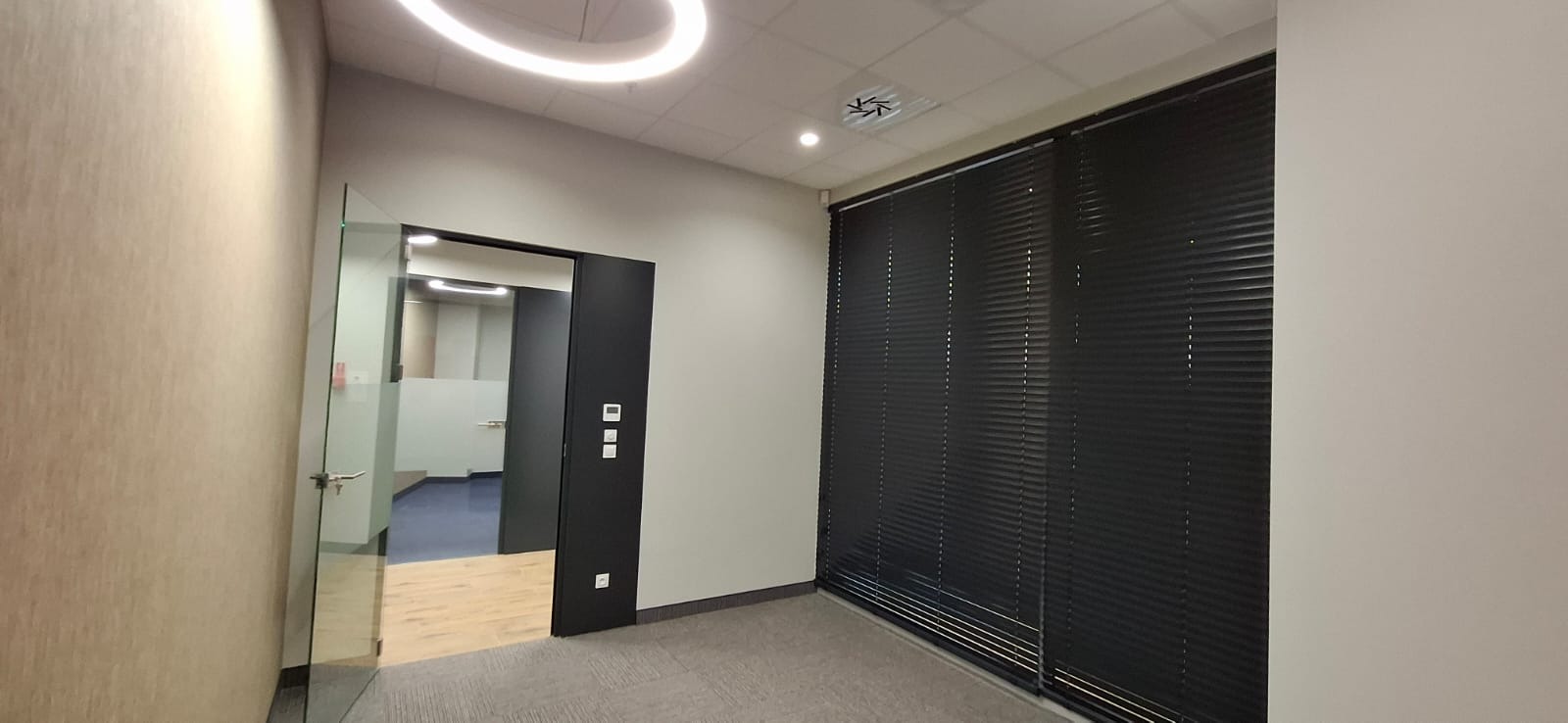 Nowoczesne biuro po remoncie: szklane drzwi, czarne żaluzje, oświetlenie LED. Minimalistyczny design, jasne ściany, szara wykładzina. Widok na kolejne pomieszczenie.