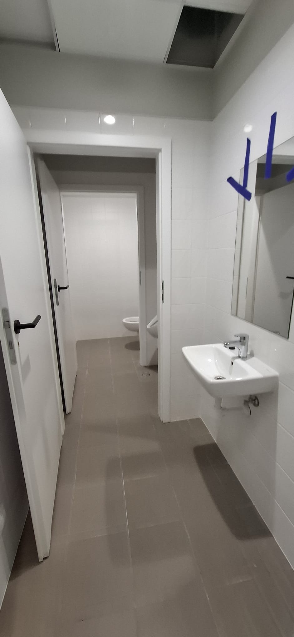 Wnętrze łazienki z umywalką, lustrem i otwartymi drzwiami do toalety. Widoczne szare płytki podłogowe i białe ściany. Minimalistyczny design.